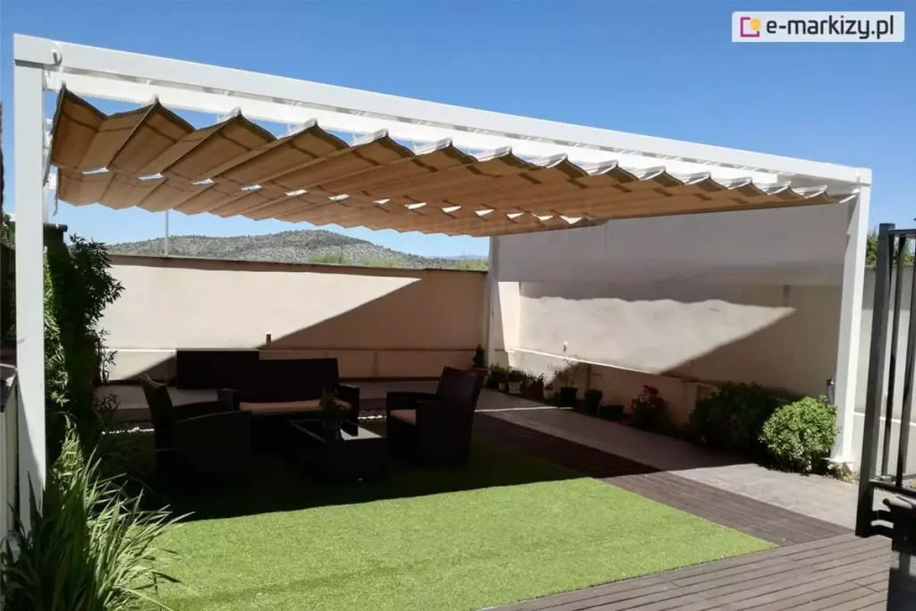 Nowoczesna, wolnostojąca pergola tarasowa o konstrukcji aluminiowej w kolorze białym, zainstalowana na prywatnej posesji. Zadaszenie stanowi system rzymski wykonany z tkaniny w kolorze jasnobrązowym/cappuccino, który jest w pełni rozłożony, tworząc regularne, poziome fale podtrzymywane przez białe profile poprzeczne. Pod pergolą zaaranżowano strefę relaksu z meblami ogrodowymi z technorattanu w kolorze ciemnego brązu (stolik kawowy, fotele i sofa) ustawionymi na fragmencie sztucznej trawy i pogrążonych w głębokim cieniu. Reszta podłoża wykonana jest z ciemnych desek kompozytowych oraz jasnych płyt tarasowych. W tle widoczny jest jasny mur odgradzający posesję, niskie nasadzenia roślinne w doniczkach oraz zarys wzgórz pod bezchmurnym, błękitnym niebem. Konstrukcja charakteryzuje się prostą, minimalistyczną formą z wyraźnie widocznymi pionowymi słupami wsporczymi i szeroką ramą górną.
