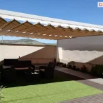 Nowoczesna, wolnostojąca pergola tarasowa o konstrukcji aluminiowej w kolorze białym, zainstalowana na prywatnej posesji. Zadaszenie stanowi system rzymski wykonany z tkaniny w kolorze jasnobrązowym/cappuccino, który jest w pełni rozłożony, tworząc regularne, poziome fale podtrzymywane przez białe profile poprzeczne. Pod pergolą zaaranżowano strefę relaksu z meblami ogrodowymi z technorattanu w kolorze ciemnego brązu (stolik kawowy, fotele i sofa) ustawionymi na fragmencie sztucznej trawy i pogrążonych w głębokim cieniu. Reszta podłoża wykonana jest z ciemnych desek kompozytowych oraz jasnych płyt tarasowych. W tle widoczny jest jasny mur odgradzający posesję, niskie nasadzenia roślinne w doniczkach oraz zarys wzgórz pod bezchmurnym, błękitnym niebem. Konstrukcja charakteryzuje się prostą, minimalistyczną formą z wyraźnie widocznymi pionowymi słupami wsporczymi i szeroką ramą górną.