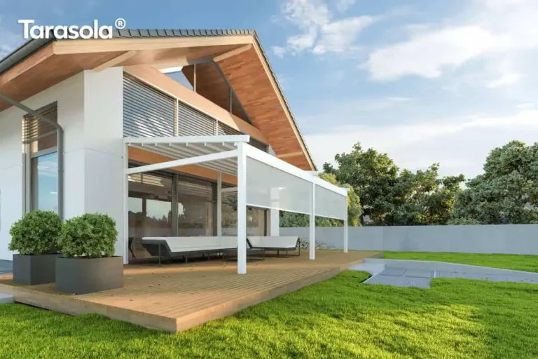 Nowoczesna, biała pergola tarasowa model Elegancy Tarasola o konstrukcji aluminiowej przyściennej, wyposażona w rzymskie, składane zadaszenie tkaninowe oraz dwie przednie rolety typu screen w kolorze jasnoszarym. Rolety są częściowo rozwinięte, co obrazuje system ochrony przed słońcem, a cała konstrukcja opiera się na stabilnych, kwadratowych profilach pionowych. Pergola została zainstalowana na przestronnym tarasie wyłożonym deskami drewnianymi w naturalnym odcieniu, na którym ustawiono duży, narożny zestaw mebli wypoczynkowych w kolorze białym. Budynek wyróżnia się współczesną architekturą z białymi ścianami, dużymi przeszkleniami wyposażonymi w srebrne żaluzje fasadowe oraz efektownym zadaszeniem z szerokim, drewnianym okapem o widocznej strukturze ziarna. W aranżacji tarasu wykorzystano duże, grafitowe donice o geometrycznym kształcie z zielonymi krzewami, a otoczenie posesji stanowi zadbany trawnik, jasny mur ogrodzeniowy oraz ściana drzew liściastych pod błękitnym niebem.