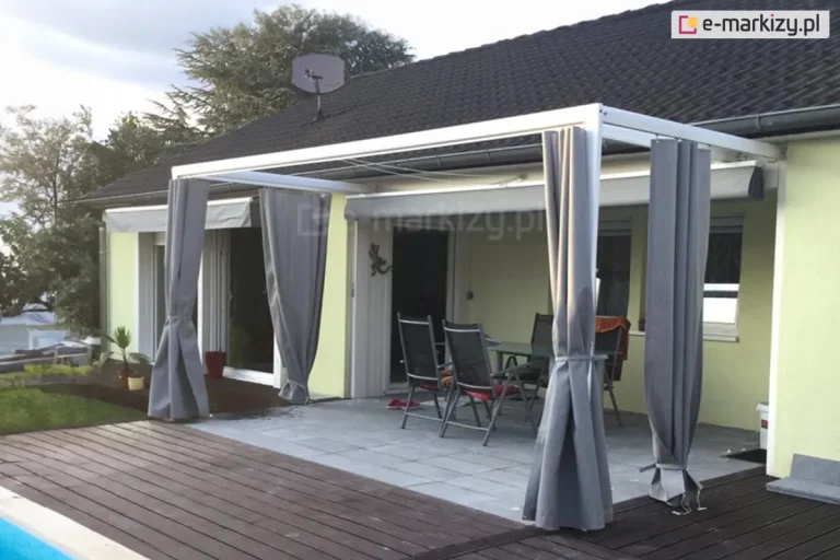 Przyścienna pergola tarasowa o konstrukcji aluminiowej w kolorze białym, zainstalowaną nad przydomowym tarasem. System wyposażony jest w dodatkowe ruchome zadaszenie tkaninowe Silva, które na zdjęciu jest całkowicie zwinięte przy elewacji. Charakterystycznym elementem tej realizacji są boczne osłony w formie zasłon ogrodowych w kolorze szarym, które są podwiązane do słupów nośnych pergoli. Taras składa się z dwóch stref: części wyłożonej szarymi płytami betonowymi, na której stoi stół z krzesłami, oraz części wykonanej z ciemnych desek kompozytowych graniczących z basenem. Budynek posiada elewację w kolorze jasnożółtym i ciemny skośny dach z dachówki. W tle widoczna jest gęsta roślinność ogrodowa oraz drzewa pod zachmurzonym niebem.