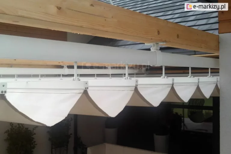 techniczne zbliżenie na system prowadnic białego dachu rzymskiego Silva, zamontowanego pod konstrukcją z naturalnego drewna. Widoczna jest główna, okrągła rura nośna w kolorze białym, która jest przytwierdzona do poziomej, jasnej belki drewnianej za pomocą specjalnych uchwytów montażowych. Od rury odchodzą pionowe trzpienie wspierające poprzeczne profile, na których rozpięte jest jasne poszycie tkaninowe. Tkanina układa się w symetryczne, trójkątne fałdy, tworząc charakterystyczny efekt dachu rzymskiego. W tle widoczny jest fragment dachu budynku z ciemnoszarą dachówką oraz zarys tarasu z roślinnością doniczkową. Perspektywa z dołu pozwala na dokładne zaobserwowanie stabilności połączeń mechanicznych oraz sposobu podwieszenia materiału.