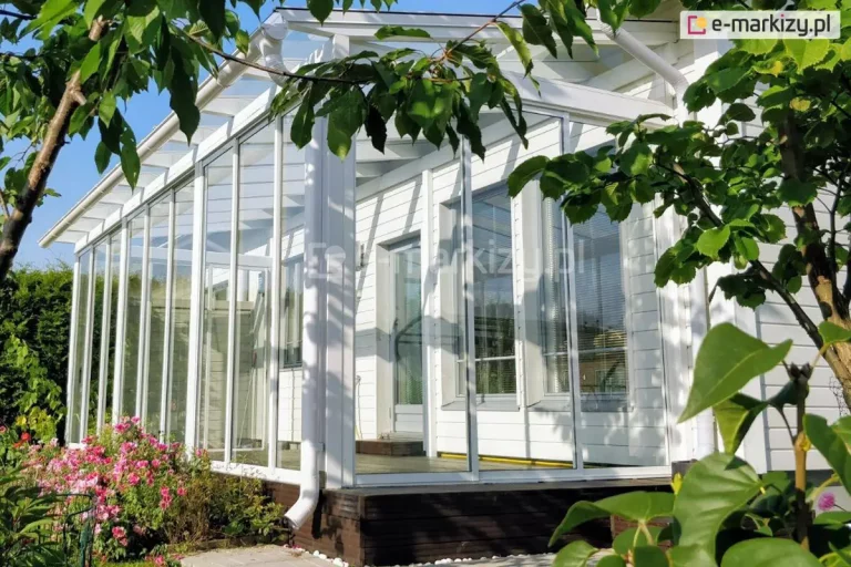 Biała, przeszklona zabudowa tarasu przylegająca do drewnianego domu o białych elewacjach. System składa się z wysokich, ramowych paneli szklanych Sotro, które są osadzone w białych profilach aluminiowych górnych i dolnych. Konstrukcja posiada jednospadowy, przezroczysty dach z widocznym systemem rynnowym odprowadzającym wodę. Zabudowa jest zamontowana na ciemnobrązowym, drewnianym podeście tarasowym. Wokół obiektu widoczna jest gęsta roślinność ogrodowa, w tym kwitnące różowe krzewy oraz liście drzew w pierwszym planie, co podkreśla integrację konstrukcji z naturą.