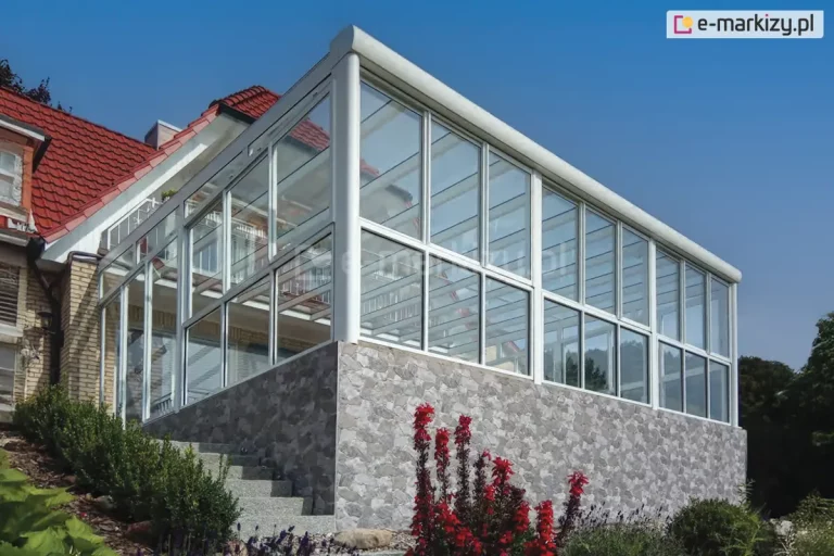 Bryła nowoczesnej, białej zabudowy szklanej zamontowanej na wysokim fundamencie obłożonym łupanym kamieniem w odcieniach szarości. Konstrukcja posiada pochylony, przeszklony dach wsparty na solidnych, okrągłych słupach narożnych w kolorze białym. Ściany boczne wykonane są w systemie Sotro oraz Fest, gdzie stałe przeszklenia są podzielone szprosami na prostokątne moduły, tworząc estetyczną i stabilną strukturę. Zabudowa jest zintegrowana z budynkiem mieszkalnym o czerwonym dachu, a wokół znajdują się zadbane nasadzenia ogrodowe, w tym czerwone kwiaty i zielone krzewy, na tle czystego, niebieskiego nieba.