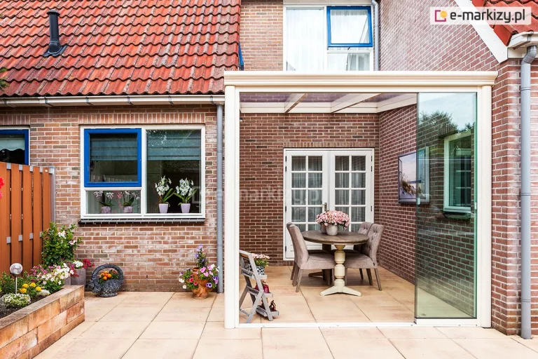 Kremowa zabudowa tarasu w formie ogrodu zimowego, zrealizowaną w narożniku piętrowego budynku z czerwonej cegły. Konstrukcja zadaszenia wykonana jest z profili aluminiowych, z dachem pokrytym przezroczystymi panelami. Frontowa część zabudowy posiada system szklanych ścian przesuwnych, z których jeden panel jest całkowicie zsunięty na bok, otwierając przejście na zewnętrzną część tarasu wyłożoną jasnymi płytami. Wewnątrz osłoniętej przestrzeni znajduje się kącik jadalniany z okrągłym, drewnianym stołem na dekoracyjnej nodze oraz czterema eleganckimi, szarymi krzesłami z pikowaniem. Na stole ustawiono donicę z różowymi kwiatami, a na ceglanej ścianie wewnątrz zabudowy powieszono obraz. W tle widoczne są białe, przeszklone drzwi prowadzące do domu. Po lewej stronie zdjęcia znajduje się fragment ogrodu z murowaną donicą pełną kwitnących kwiatów oraz okno z niebieską ramą, na którego parapecie stoją doniczki ze storczykami.