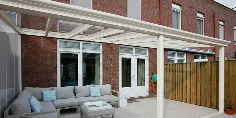 Białe aluminiowe zadaszenie tarasowe Pigato marki Deponti (nazwa handlowa Terano), o klasycznym designie, zamontowane do elewacji budynku wykonanej z czerwonej cegły. Konstrukcja zadaszenia opiera się na przyściennej belce nośnej oraz trzech (widoczne są tylko dwa) frontowych słupach o zaokrąglonym profilu, wspierających system rynnowy. Dach zadaszenia jest przeszklony, wykonany z transparentnych paneli podzielonych białymi profilami dachowymi, co zapewnia pełny dostęp światła dziennego. Pod zadaszeniem zaaranżowano strefę wypoczynkową z dużą, narożną sofą w kolorze szarym, udekorowaną błękitnymi poduszkami, oraz pasującym pufem pełniącym funkcję stolika. W tle widoczne są białe drzwi tarasowe i okna budynku, a po prawej stronie wysokie ogrodzenie z pionowych drewnianych desek. Taras wyłożony jest dużymi, jasnoszarymi płytami, a z lewego boku zamontowano przezroczysty panel boczny, wykonany z poliwęglanu komorowego, chroniący przed wiatrem.