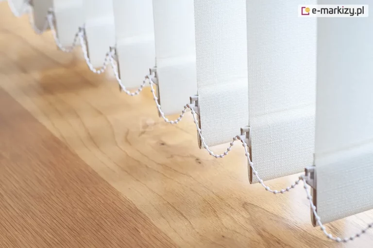 Dolna część białych żaluzji pionowych wiszących tuż nad jasną drewnianą podłogą o wyraźnym usłojeniu. Na zdjęciu widocznych jest kilka pionowych pasów materiału o drobnej, tekstylnej strukturze, które są ustawione pod skosem względem osi okna. Każdy pas żaluzji pionowej posiada na dole wszyte obciążniki ukryte w materiale, które zakończone są plastikowymi uszkami po obu stronach. Przez te uszka przeciągnięty jest biały łańcuszek kulkowy, który łączy poszczególne lamele, zapewniając ich równomierne rozkładanie się oraz stabilność podczas przesuwania. Łańcuszek układa się w charakterystyczne, miękkie łuki między kolejnymi pasami tkaniny. Światło padające z boku podkreśla splot materiału żaluzji oraz gładką powierzchnię lakierowanej podłogi, tworząc delikatne cienie. Perspektywa zdjęcia jest ujęta pod kątem, co pozwala dostrzec głębię i powtarzalność elementów składowych systemu osłonowego.