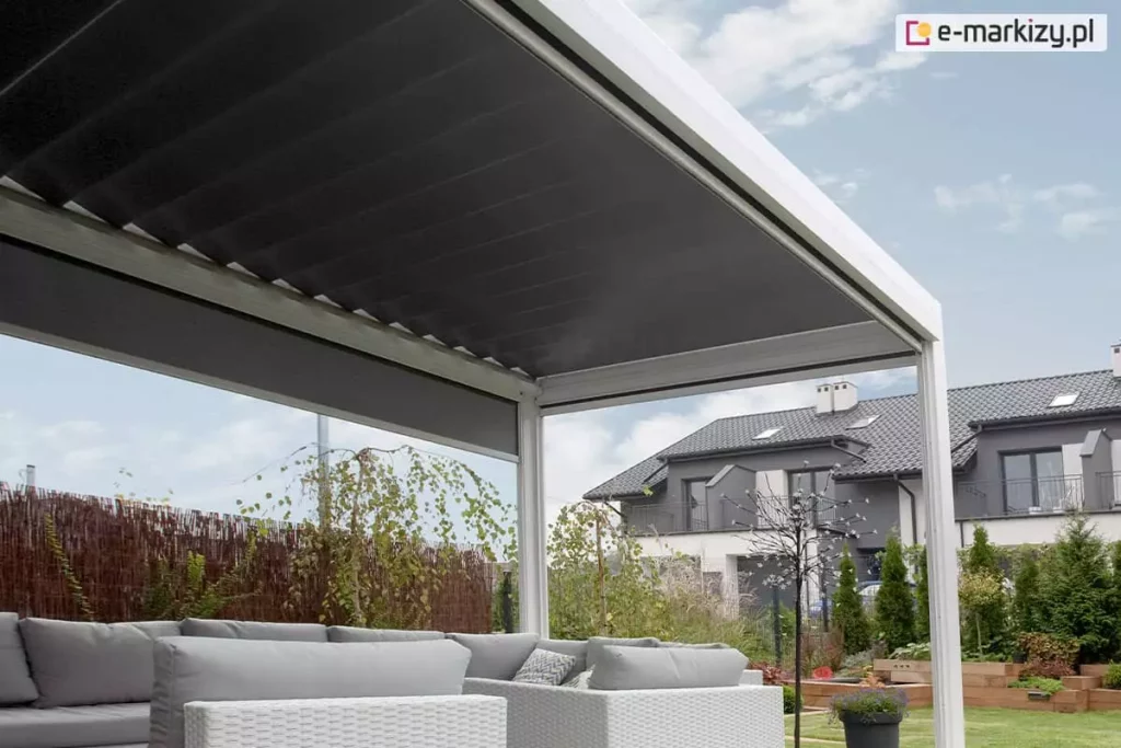 Pergola tarasowa Sb400 firmy Aluprof w kolorze białym wyposażona w zewnętrzną roletę boczną nas jasnymi meblami ogrodowymi, całość doskonale podkreśla piękną zieloną roślinność w ogrodzie. W tle widoczny także sąsiedni budynek szeregowy.