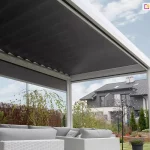Pergola tarasowa Sb400 firmy Aluprof w kolorze białym wyposażona w zewnętrzną roletę boczną nas jasnymi meblami ogrodowymi, całość doskonale podkreśla piękną zieloną roślinność w ogrodzie. W tle widoczny także sąsiedni budynek szeregowy.