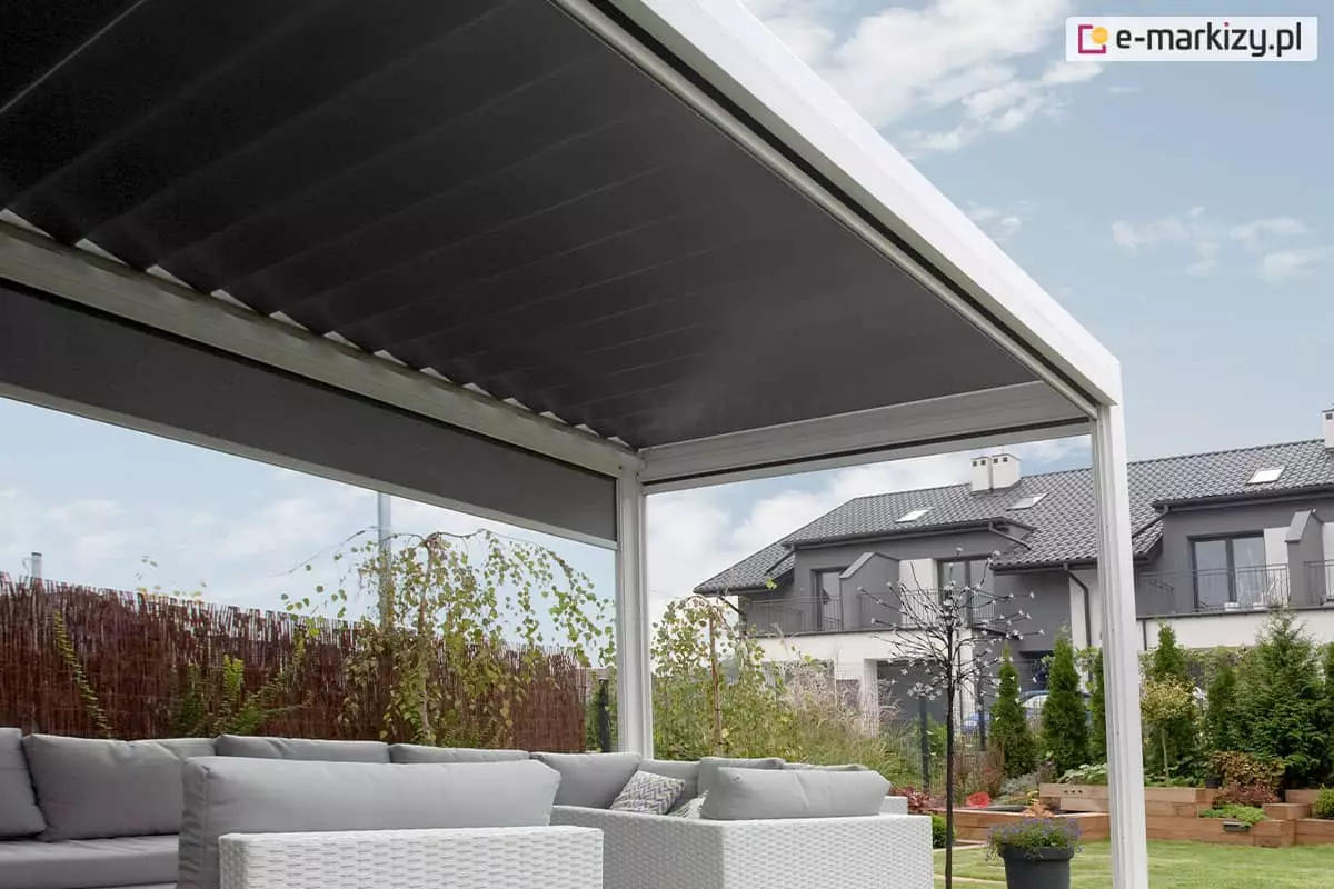 Pergola tarasowa Sb400 firmy Aluprof w kolorze białym wyposażona w zewnętrzną roletę boczną nas jasnymi meblami ogrodowymi, całość doskonale podkreśla piękną zieloną roślinność w ogrodzie. W tle widoczny także sąsiedni budynek szeregowy.
