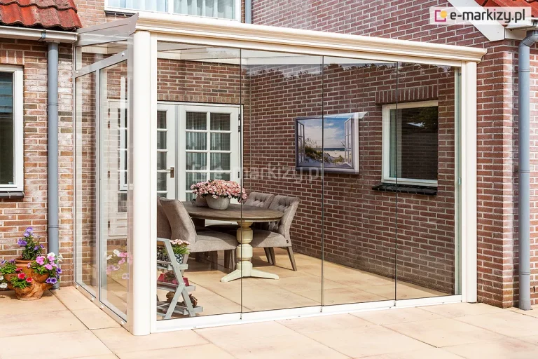 Narożne zadaszenie tarasowe w formie ogrodu zimowego, o konstrukcji aluminiowej w kolorze kremowym, zamontowane we wnęce budynku z czerwonej cegły. Konstrukcja posiada szklane ściany przesuwne, które całkowicie zamykają przestrzeń, oraz przezroczysty dach osadzony na profilach aluminiowych. Wewnątrz pomieszczenia znajduje się strefa jadalniana z owalnym, drewnianym stołem na dekoracyjnej nodze oraz czterema tapicerowanymi krzesłami w kolorze beżowym. Na stole ustawiona jest duża donica z różowymi kwiatami. Podłoga wewnątrz i na zewnątrz wykonana jest z jednolitych, jasnych płyt tarasowych o dużym formacie. Na elewacji budynku, w obrębie zabudowy, widoczne są białe drzwi z przeszkleniami oraz okno, a na ścianie zawieszono obraz przedstawiający nadmorski krajobraz. Po lewej stronie, przed szklaną ścianą, stoi dekoracyjna donica z kolorowymi kwiatami oraz mały, drewniany kwietnik w formie drabinki.