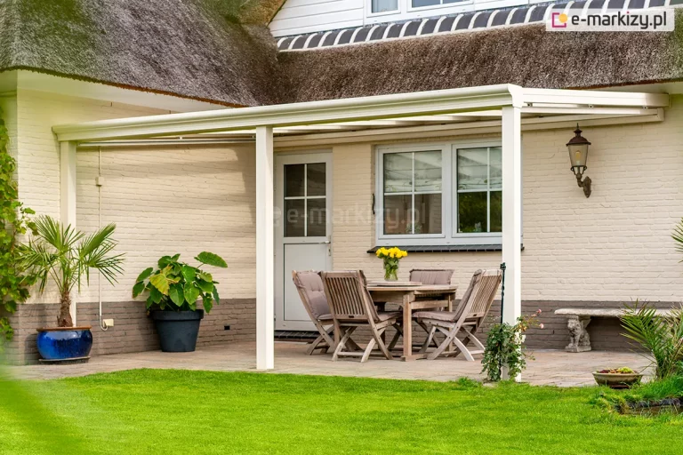 Eleganckie zadaszenie tarasowe Terano w kolorze białym, zamontowane przy budynku o jasnej, ceglanej elewacji i dachu pokrytym naturalną strzechą. Aluminiowa konstrukcja o minimalistycznym designie składa się z ramy głównej, krokwi dachowych oraz pionowych słupów nośnych o kwadratowym przekroju. Pod zadaszeniem zaaranżowano klasyczną strefę wypoczynkową z drewnianym stołem i dopasowanymi krzesłami ogrodowymi z miękkimi poduszkami w kolorze beżowym. Na tarasie znajdują się również rośliny egzotyczne w dekoracyjnych donicach, w tym palma, oraz stylowa lampa ścienna w formie latarni. Przestrzeń tarasu płynnie łączy się z zadbanym, zielonym trawnikiem. Konstrukcja dachu posiada wypełnienie ze szkła, zapewniające ochronę przed opadami przy zachowaniu dostępu do światła dziennego.