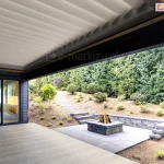 nowoczesny taras z dachem lamelowym Selt SB400R w kontrastowym zestawieniu kolorystycznym. Lamele dachu są w kolorze białym i znajdują się w pozycji lekko uchylonej, zapewniając swobodny przepływ powietrza na tarasie. Lamele w pozycji zamkniętej tworzą szczelną, gładką płaszczyznę chroniącą przed opadami. Rama okalająca zadaszenie oraz konstrukcja budynku są wykonane w kolorze ciemnego grafitu. Taras posiada podłogę z jasnych desek kompozytowych i graniczy bezpośrednio z ogrodem o zróżnicowanym ukształtowaniu terenu. Na zewnątrz widoczne jest palenisko ogrodowe obudowane kamieniem, schody terenowe oraz gęsta roślinność iglasta. Przez duże przeszklenie z boku widać wnętrze salonu z ciepłym oświetleniem, co podkreśla integrację strefy domowej z wypoczynkową częścią zewnętrzną.