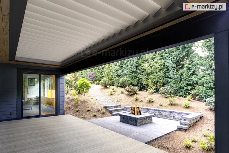nowoczesny taras z dachem lamelowym Selt SB400R w kontrastowym zestawieniu kolorystycznym. Lamele dachu są w kolorze białym i znajdują się w pozycji lekko uchylonej, zapewniając swobodny przepływ powietrza na tarasie. Lamele w pozycji zamkniętej tworzą szczelną, gładką płaszczyznę chroniącą przed opadami. Rama okalająca zadaszenie oraz konstrukcja budynku są wykonane w kolorze ciemnego grafitu. Taras posiada podłogę z jasnych desek kompozytowych i graniczy bezpośrednio z ogrodem o zróżnicowanym ukształtowaniu terenu. Na zewnątrz widoczne jest palenisko ogrodowe obudowane kamieniem, schody terenowe oraz gęsta roślinność iglasta. Przez duże przeszklenie z boku widać wnętrze salonu z ciepłym oświetleniem, co podkreśla integrację strefy domowej z wypoczynkową częścią zewnętrzną.