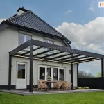 Nowoczesna i funkcjonalna zabudowa tarasu Spekta Glass ujęta z dalszej perspektywy, co pozwala na szczegółową ekspozycję konstrukcji dachu oraz aranżacji przestrzeni wypoczynkowej. System został zintegrowany z bryłą budynku o białej elewacji i skośnym dachu krytym szarą blachą, tworząc estetyczne przedłużenie salonu. Rama nośna oraz słupy wsporcze o przekroju kwadratowym wykonane są z profili aluminiowych w kolorze antracytowym. Zadaszenie jest w całości przeszklone i podzielone na kilka regularnych sekcji za pomocą podłużnych krokwi, co zapewnia pełną ochronę przed opadami przy jednoczesnym zachowaniu maksymalnej jasności na tarasie. Pod konstrukcją zaaranżowano strefę relaksu wyposażoną w meble z technorattanu w naturalnym odcieniu, w tym dwie sofy z szarymi poduchami, fotele oraz pufy. Podłoże tarasu stanowią wielkoformatowe płyty ceramiczne w kolorze jasnoszarym, które sąsiadują z zadbanym trawnikiem. Całość aranżacji dopełniają liczne rośliny ozdobne w białych donicach, podkreślające nowoczesny i harmonijny charakter realizacji.