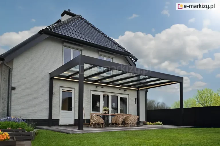 Nowoczesna i funkcjonalna zabudowa tarasu Spekta Glass ujęta z dalszej perspektywy, co pozwala na szczegółową ekspozycję konstrukcji dachu oraz aranżacji przestrzeni wypoczynkowej. System został zintegrowany z bryłą budynku o białej elewacji i skośnym dachu krytym szarą blachą, tworząc estetyczne przedłużenie salonu. Rama nośna oraz słupy wsporcze o przekroju kwadratowym wykonane są z profili aluminiowych w kolorze antracytowym. Zadaszenie jest w całości przeszklone i podzielone na kilka regularnych sekcji za pomocą podłużnych krokwi, co zapewnia pełną ochronę przed opadami przy jednoczesnym zachowaniu maksymalnej jasności na tarasie. Pod konstrukcją zaaranżowano strefę relaksu wyposażoną w meble z technorattanu w naturalnym odcieniu, w tym dwie sofy z szarymi poduchami, fotele oraz pufy. Podłoże tarasu stanowią wielkoformatowe płyty ceramiczne w kolorze jasnoszarym, które sąsiadują z zadbanym trawnikiem. Całość aranżacji dopełniają liczne rośliny ozdobne w białych donicach, podkreślające nowoczesny i harmonijny charakter realizacji.