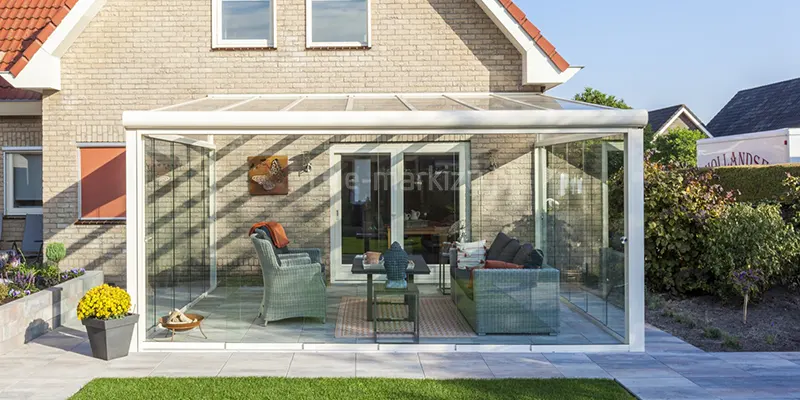 Zdjęcie prezentuje biały, w pełni przeszklony ogród zimowy oparty na systemie Terano, zamontowany do elewacji budynku z jasnej cegły. Konstrukcja posiada dach z przezroczystych paneli oraz ściany frontowe i boczne wykonane z bezramowych, przesuwnych tafli szkła hartowanego, które są osadzone w białych szynach prowadzących. Pod zadaszeniem zaaranżowano komfortową strefę wypoczynkową z meblami z szarego technorattanu, w tym sofę z ciemnymi poduszkami oraz dwa wysokie fotele, ustawione wokół czarnego stolika kawowego. Wewnątrz znajduje się również dekoracyjny dywan w geometryczne wzory, małe palenisko oraz ozdobne figurki. Taras wyłożony jest jasnoszarymi płytami, a przed nim rozciąga się zielony trawnik. Po lewej stronie widoczna jest murowana donica z żółtymi kwiatami, a w tle gęsta roślinność ogrodowa oraz fragment białego pojazdu ciężarowego. Słońce rzuca wyraźne, ukośne cienie konstrukcji na elewację budynku i wnętrze tarasu.