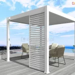 Biała wolnostojąca pergola lamelowa Marlo o kwadratowej podstawie, zamontowana na tarasie z widokiem na morze. Konstrukcja zadaszenia posiada dach z ruchomych lamel ustawionych w pozycji całkowicie zamkniętej oraz pionowy, biały panel żaluzjowy zamontowany w jednym z narożników, który służy jako przesłona boczna. Przy słupie nośnym po lewej stronie widoczny jest mechanizm korbowy do ręcznego sterowania kątem nachylenia lamel dachowych. Pod pergolą, na podłożu z jasnoszarych desek tarasowych, ustawiono dwa nowoczesne fotele z ażurowym splotem i jasnymi poduszkami. Taras zabezpieczony jest niską, białą barierką z przeszkleniami, za którą rozpościera się widok na wybrzeże i błękitną wodę pod jasnym niebem z delikatnymi chmurami.