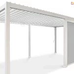 Wolnostojąca pergola bioklimatyczna Gloria o konstrukcji modułowej wykonana z aluminium w kolorze białym przedstawiona na jednolitym jasnym tle. Produkt składa się z dwóch połączonych segmentów wspartych na sześciu pionowych słupach o kwadratowym przekroju z widocznymi stopami montażowymi. Zadaszenie tworzy system poziomych ruchomych lameli aluminiowych w kolorze białym, które są ustawione w pozycji otwartej. Jeden z bocznych modułów pergoli został wyposażony w zintegrowaną roletę pionową typu screen w kolorze jasnoszarym, która jest całkowicie rozwinięta i osłania przestrzeń między dwoma słupami. Przy jednym ze słupów widoczna jest metalowa korba służąca do manualnej regulacji kąta nachylenia lameli dachowych. Całość konstrukcji charakteryzuje się minimalistycznym, nowoczesnym designem z ukrytymi systemami odprowadzania wody wewnątrz profili.
