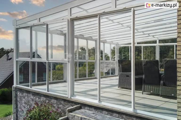 Fotografia ukazuje rozbudowaną zabudowę tarasu na podwyższeniu, pełniącą funkcję ogrodu zimowego, wykonaną w systemie białych profili aluminiowych. Głównym elementem są wieloskrzydłowe drzwi przesuwne Sotro, które pozwalają na otwarcie szerokich przejść. Konstrukcja posiada przezroczyste zadaszenie oraz stałe przeszklenia boczne, co maksymalizuje doświetlenie wnętrza. Taras jest posadowiony na murku wykończonym szarym kamieniem dekoracyjnym, a w środku znajduje się zestaw wypoczynkowy z ciemnego technorattanu. W tle widoczna jest panorama z budynkami mieszkalnymi oraz drzewami pod niebem w barwach zachodzącego słońca.