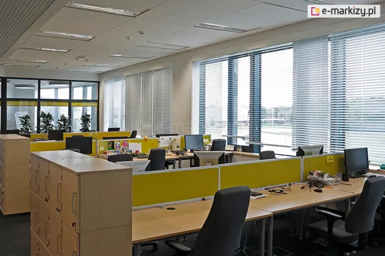 Widok na nowoczesne biuro typu open space z szerokim przeszkleniem wyposażonym w wewnętrzne żaluzje fasadowe C50 Slim w kolorze srebrnym. Lamele żaluzji są ustawione w pozycji otwartej, co pozwala na precyzyjną kontrolę dopływu światła dziennego i redukcję odblasków na monitorach, przy jednoczesnym zachowaniu widoku na zewnętrzną infrastrukturę. Wnętrze podzielone jest na stanowiska pracy za pomocą żółtych paneli biurkowych, które pełnią funkcję przegród akustycznych i wizualnych. Wyposażenie biura obejmuje jasne biurka, ergonomiczne czarne fotele oraz regały z szafkami na dokumenty w kolorze naturalnego drewna. Na suficie kasetonowym widoczne są wbudowane oprawy oświetleniowe oraz czujniki systemów budynkowych, a cała aranżacja podkreśla profesjonalny i funkcjonalny charakter przestrzeni biurowej wykorzystującej nowoczesne systemy osłonowe.