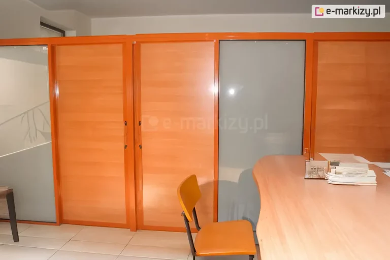 Zdjęcie ukazuje zabudowę biurową wykonaną z pomarańczowych profili aluminiowych, stanowiącą system ścian stałych Fest. Przegrody posiadają mieszane wypełnienie: panele o strukturze jasnego drewna imitujące boazerię oraz tafle matowego, mlecznego szkła. Na pierwszym planie widoczne jest biurko z jasnym blatem oraz krzesło z pomarańczowym siedziskiem, które kolorystycznie nawiązuje do ram konstrukcyjnych.