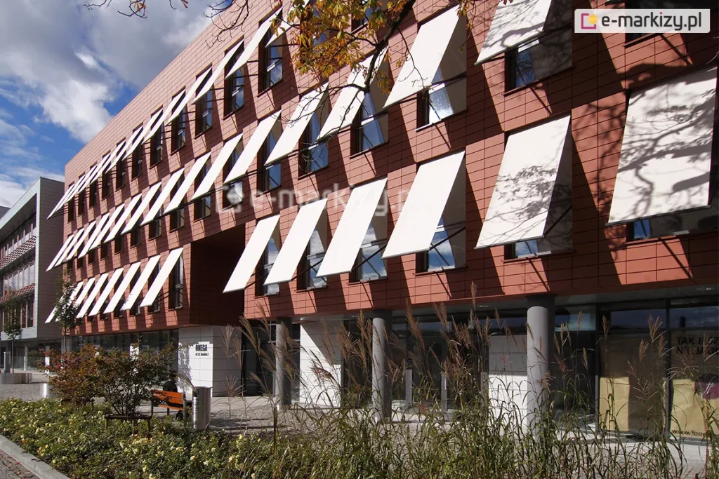 Elewacja nowoczesnego budynku biurowego, wykończona ceramicznymi płytami w kolorze ceglastym, z rzędami pionowych okien wyposażonych w system zewnętrznych osłon przeciwsłonecznych. Na trzech kondygnacjach obiektu zamontowano liczne markizy sklepowe Italia Aluprof z tkaniną akrylową w jasnym, niemal białym kolorze, które są wysunięte pod kątem, tworząc powtarzalny, geometryczny wzór na fasadzie. Parter budynku jest przeszklony i cofnięty względem lica wyższych pięter, wsparty na szarych, cylindrycznych słupach konstrukcyjnych. Przed budynkiem znajduje się zagospodarowany teren z niskimi krzewami, ozdobnymi trawami oraz elementami małej architektury, takimi jak metalowy kosz na śmieci i drewniana ławka. W prawej części kadru na jasnej tkaninie markiz widoczne są cienie rzucane przez gałęzie pobliskiego drzewa. Niebo jest częściowo zachmurzone, a naturalne oświetlenie podkreśla kontrast między ciepłym odcieniem elewacji a jasnymi płaszczyznami rozwiniętych osłon.