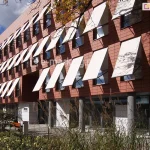 Elewacja nowoczesnego budynku biurowego, wykończona ceramicznymi płytami w kolorze ceglastym, z rzędami pionowych okien wyposażonych w system zewnętrznych osłon przeciwsłonecznych. Na trzech kondygnacjach obiektu zamontowano liczne markizy sklepowe Italia Aluprof z tkaniną akrylową w jasnym, niemal białym kolorze, które są wysunięte pod kątem, tworząc powtarzalny, geometryczny wzór na fasadzie. Parter budynku jest przeszklony i cofnięty względem lica wyższych pięter, wsparty na szarych, cylindrycznych słupach konstrukcyjnych. Przed budynkiem znajduje się zagospodarowany teren z niskimi krzewami, ozdobnymi trawami oraz elementami małej architektury, takimi jak metalowy kosz na śmieci i drewniana ławka. W prawej części kadru na jasnej tkaninie markiz widoczne są cienie rzucane przez gałęzie pobliskiego drzewa. Niebo jest częściowo zachmurzone, a naturalne oświetlenie podkreśla kontrast między ciepłym odcieniem elewacji a jasnymi płaszczyznami rozwiniętych osłon.