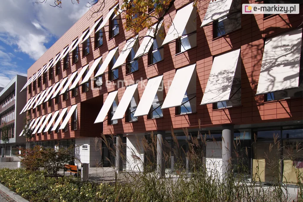 Elewacja nowoczesnego budynku biurowego, wykończona ceramicznymi płytami w kolorze ceglastym, z rzędami pionowych okien wyposażonych w system zewnętrznych osłon przeciwsłonecznych. Na trzech kondygnacjach obiektu zamontowano liczne markizy sklepowe Italia z tkaniną akrylową w jasnym, niemal białym kolorze, które są wysunięte pod kątem, tworząc powtarzalny, geometryczny wzór na fasadzie. Parter budynku jest przeszklony i cofnięty względem lica wyższych pięter, wsparty na szarych, cylindrycznych słupach konstrukcyjnych. Przed budynkiem znajduje się zagospodarowany teren z niskimi krzewami, ozdobnymi trawami oraz elementami małej architektury, takimi jak metalowy kosz na śmieci i drewniana ławka. W prawej części kadru na jasnej tkaninie markiz widoczne są cienie rzucane przez gałęzie pobliskiego drzewa. Niebo jest częściowo zachmurzone, a naturalne oświetlenie podkreśla kontrast między ciepłym odcieniem elewacji a jasnymi płaszczyznami rozwiniętych osłon.