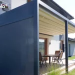 Zdjęcie prezentuje pergolę przyścienną Tarasola Flat o antracytowej konstrukcji aluminiowej, zamontowaną przy białej elewacji budynku jednorodzinnego. Konstrukcja wspiera się na trzech równomiernie rozłożonych słupach podporowych o kwadratowym przekroju. Model ten posiada dach tkaninowy w kolorze kremowym, który jest w pełni rozwinięty i wyposażony w zintegrowane, punktowe oświetlenie LED rozmieszczone na profilach poprzecznych. Charakterystycznym elementem tej realizacji jest boczna osłona typu screen (zip) w kolorze ciemnoszarym, która jest całkowicie opuszczona, zamykając jeden z boków pergoli i zapewniając ochronę przed wiatrem oraz słońcem. Pod zadaszeniem, na tarasie wyłożonym jasnobrązowymi płytkami, ustawiono drewniany stół z krzesłami. W tle widoczny jest zielony trawnik, niskie nasadzenia ozdobne oraz sąsiednie domy jednorodzinne pod czystym, błękitnym niebem.