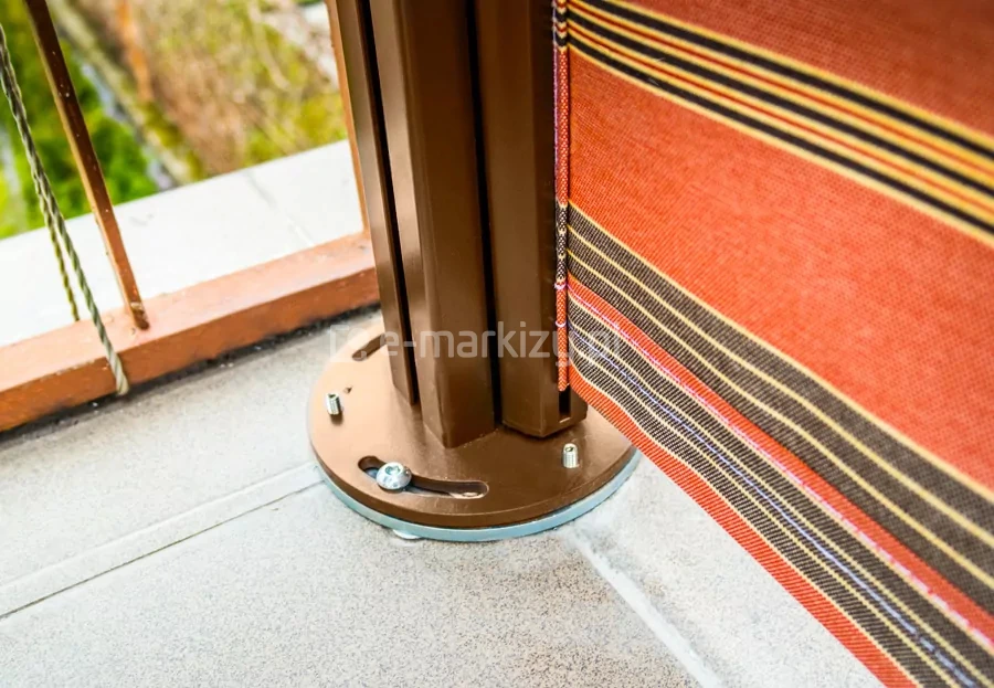 Zbliżenie mocowania słupka mocowanego do podłoża twardego - wariant mocowania typ1 , w kolorze brązowym, zamocowanego do powierzchni tarasu pokrytej płytkami gresowymi. Przy słupku widoczna belka przednia z poszyciem w pasy o dominującym kolorze czerwonym. W tle za mocowaniem barierka balkonowa w kolorze pomarańczowym.