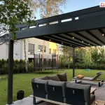 Nowoczesny ogród z tarasem osłoniętym minimalistyczną pergolą Square flat w kolorze grafitowym. Dach konstrukcji zbudowany jest z szerokich pasów materiału ze zintegrowanym oświetleniem LED – światła umieszczone w niektórych poprzeczkach są włączone. Od boku pergoli na pierwszym planie widoczna charakterystyczna rynna odpływowa, zapewniająca suchą powierzchnię pod zadaszeniem. Pod zadaszeniem ustawiono elegancki zestaw mebli ogrodowych – narożną sofę z tapicerką w kolorze jasnej szarości i drewnianą podstawą, oraz pojedynczy leżak z poduszką w kolorze musztardowym. Taras wyłożony jest dużymi, jasnoszarymi płytami. Przy sofie stoi niski, czarny stolik o otwartej, koszykowej strukturze. Tło to przestronny ogród otoczony żywopłotem i różnorodnymi drzewami liściastymi i iglastymi. W głębi widać dużą, okrągłą trampolinę z siatką zabezpieczającą, a dalej dziecięce sprzęty do zabawy – zjeżdżalnię i huśtawkę. Na lewo znajduje się sąsiedni dom z prostą, nowoczesną elewacją. Kompozycja oddaje harmonijne połączenie estetyki wypoczynku dla dorosłych i przestrzeni rekreacyjnej dla dzieci.
