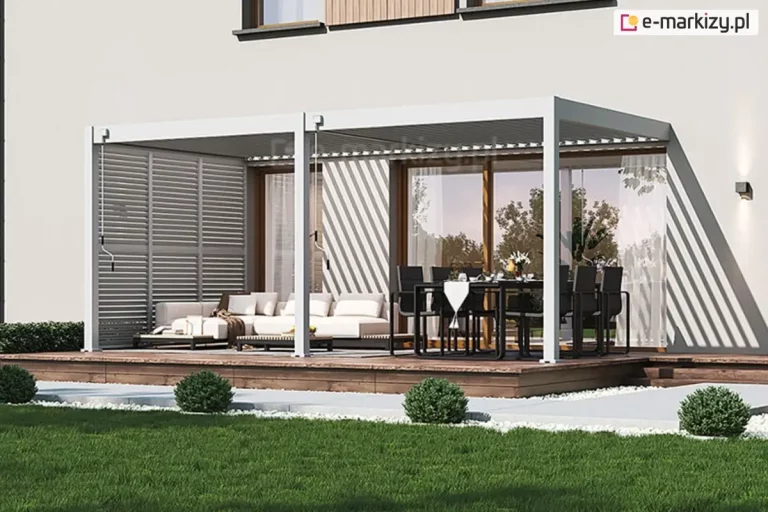 Biała bioklimatyczna pergola Marlo o konstrukcji dwumodułowej, zamontowana przy jasnej elewacji nowoczesnego budynku. Zadaszenie wsparte jest na trzech frontowych słupach aluminiowych, a dach tworzą ruchome lamele, które ustawione pod kątem, rzucają pasy cienia na ścianę domu. Lewa strona pergoli została wyposażona w pełną zabudowę boczną w formie stałych paneli żaluzjowych w kolorze białym, które osłaniają strefę wypoczynkową z jasną sofą narożną. Po prawej stronie znajduje się strefa jadalniana z czarnym stołem i sześcioma dopasowanymi krzesłami, ustawiona przed dużym przeszkleniem tarasowym. Całość konstrukcji osadzona jest na podwyższonym tarasie z naturalnych, brązowych desek, do którego prowadzą niskie stopnie. Otoczenie stanowi pas jasnego żwiru z niskimi, kulistymi krzewami oraz zadbany zielony trawnik. Przy słupach widoczne są mechanizmy korbowe do ręcznej obsługi dachu.