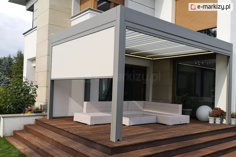 Piętrowy budynek z umieszczonym na parterze w pobliżu dwóch dużych okien drewnianym tarasem osłoniętym białą pergolą. Na tarasie ustawiono meble z rattanu w jasnym kolorze oraz rośliny doniczkowe. Na ścianach pergoli zamocowane refleksole ZIP L ZIP U, które chronią powierzchnię pod pergolą przed słońcem, deszczem i wiatrem. Drewniane stopnie prowadzą z tarasu na trawnik.