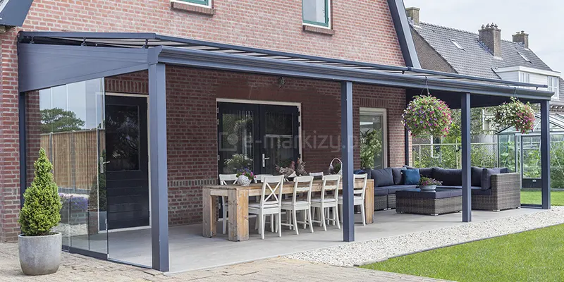 Zbliżenie na boczny element konstrukcyjny zadaszenia tarasowego model Terano, stanowiący wypełnienie przestrzeni pod skosem dachu, czyli tzw. klin boczny. Element ten składa się z aluminiowej ramy w kolorze antracytowym, w której osadzony został fragment płaskiej blachy, o trójkątnym kształcie. Poniżej klina zamontowano system szklanych drzwi przesuwnych, z których jeden panel jest częściowo otwarty. Konstrukcja zadaszenia jest zintegrowana z budynkiem o elewacji z czerwonej cegły, wyposażonym w ciemne drzwi z przeszkleniem. Na tarasie, wyłożonym jasnoszarymi płytami, znajduje się strefa jadalniana z masywnym drewnianym stołem oraz białymi krzesłami z charakterystycznym krzyżowaniem na oparciach. W tle widoczny jest fragment ogrodu z trawnikiem oraz nasadzenia w donicach, w tym formowany iglak w szarym pojemniku ustawiony na brukowanym podjeździe.