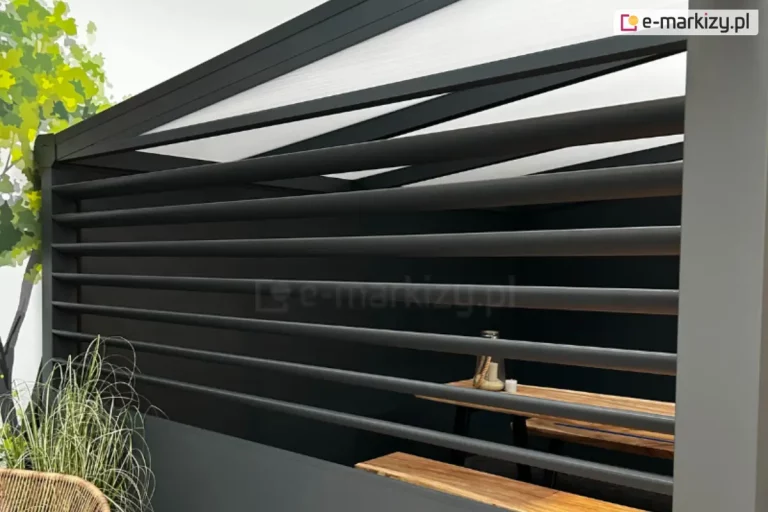 Zbliżenie na narożnik nowoczesnego zadaszenia tarasu, w którym na bocznej ścianie zamontowano aluminiowy panel żaluzjowy Louvre w kolorze głębokiego antracytu. Zdjęcie ukazuje konstrukcję z perspektywy bocznej, podkreślając precyzyjne wykończenie poziomych lameli tworzących stałą przesłonę. Nad panelem żaluzjowym Louvre widoczny jest fragment dachu z wypełnieniem z poliwęglanu komorowego w odcieniu melcznym oraz ciemnymi profilami konstrukcyjnymi ułożonymi pod skosem. Przez prześwity między lamelami żaluzji można dostrzec wnętrze strefy wypoczynkowej, w której znajduje się stół z grubym blatem z litego drewna oraz ława o podobnej stylistyce, na stole ustawiono dekoracyjną świecę w szklanym naczyniu. Po lewej stronie, na pierwszym planie, widoczna jest ozdobna, wysoka trawa o cienkich, zielonych liściach posadzona w donicy, a za nią fragment pnia i zielonych liści drzewa. Po prawej stronie kadr zamyka pionowy, masywny słup nośny zadaszenia. Całość kompozycji utrzymana jest w nowoczesnej, technicznej estetyce, łączącej surowość metalu z naturalnymi akcentami drewna i roślinności.