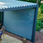 Nowoczesna pergola tarasowa o konstrukcji bioklimatycznej w kolorze antracytowym wyposażona w system ruchomych lameli dachowych oraz boczną roletę typu screen. Aluminiowa konstrukcja o ciemnych profilach została zamontowana na drewnianym podeście tarasowym w otoczeniu gęstej roślinności ogrodowej i krzewów. Zadaszenie składa się z równolegle ułożonych, jasnych lameli ustawionych pod skosem, co umożliwia regulację nasłonecznienia. Jedna z bocznych ścian pergoli jest całkowicie osłonięta szarą tkaniną techniczną rolety zewnętrznej, która chroni przed słońcem i wiatrem. Pod zadaszeniem znajduje się stół z białym obrusem oraz złote krzesła, przy których siedzą dwie kobiety w letnich ubraniach. Podłoże wokół konstrukcji wyłożone jest brązową ściółką dekoracyjną, a obok słupa nośnego stoi ozdobny lampion.