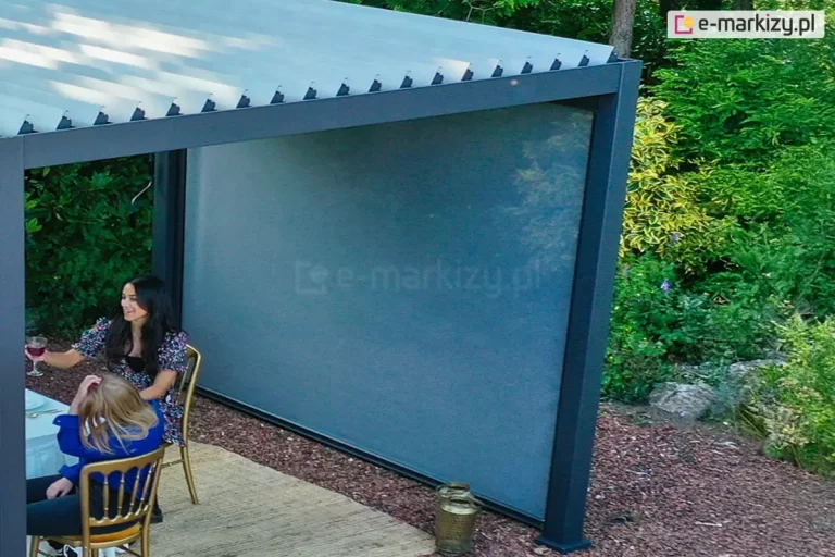 Nowoczesna pergola tarasowa o konstrukcji bioklimatycznej w kolorze antracytowym wyposażona w system ruchomych lameli dachowych oraz boczną roletę typu screen. Aluminiowa konstrukcja o ciemnych profilach została zamontowana na drewnianym podeście tarasowym w otoczeniu gęstej roślinności ogrodowej i krzewów. Zadaszenie składa się z równolegle ułożonych, jasnych lameli ustawionych pod skosem, co umożliwia regulację nasłonecznienia. Jedna z bocznych ścian pergoli jest całkowicie osłonięta szarą tkaniną techniczną rolety zewnętrznej, która chroni przed słońcem i wiatrem. Pod zadaszeniem znajduje się stół z białym obrusem oraz złote krzesła, przy których siedzą dwie kobiety w letnich ubraniach. Podłoże wokół konstrukcji wyłożone jest brązową ściółką dekoracyjną, a obok słupa nośnego stoi ozdobny lampion.