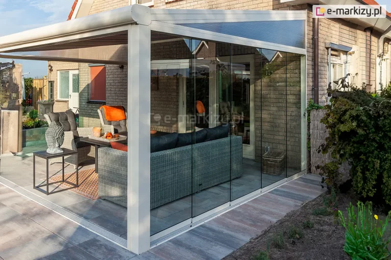 widok boczny pergoli tarasowej z zainstalowanym systemem szklanych ścian przesuwnych Auris. Konstrukcja w kolorze białym przylega do ściany domu z jasnej cegły i posiada jednospadowy dach. Panele szklane są bezramowe, połączone niemal niewidocznymi uszczelkami pionowymi, co zapewnia pełną transparentność. Przestrzeń pomiędzy skośnym dachem a bocznymi panelami szklanymi wypełniono trójkątami z poliwęglanu komorowego. Wewnątrz pergoli widoczna jest strefa wypoczynkowa z szarą sofą narożną z technorattanu, stolikiem kawowym z dekoracyjną figurą Buddy oraz fotelami. Podłoże stanowią duże, prostokątne płyty tarasowe w odcieniach szarości i brązu, a obok konstrukcji znajduje się pas ziemi przygotowany pod nasadzenia oraz fragmenty zielonej roślinności.
