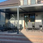 Zdjęcie przedstawia gotową realizację dużej, dwumodułowej pergoli tarasowej zamontowanej przy nowoczesnym domu jednorodzinnym. Konstrukcja wykonana jest z aluminium w kolorze ciemnego antracytu i przylega bezpośrednio do jasnej elewacji budynku, pod balkonem z metalową balustradą. Pergola posiada płaski dach z ruchomych lameli, które są w pozycji zamkniętej. Boczne ściany zabudowy zostały wyposażony w system paneli bocznych z poziomymi, ruchomymi lamelami, które tworzą ażurową ścianę osłaniającą strefę wypoczynkową. Pod zadaszeniem zaaranżowano komfortową przestrzeń relaksu, na którą składają się ciemnoszary zestaw mebli modułowych z grubymi poduszkami ustawiony w narożniku, wiszący fotel typu kokon w kolorze szarym zawieszony na własnym stelażu oraz strefa jadalniana z dużym, ciemnym stołem i czterema dopasowanymi kolorystycznie krzesłami o wysokich oparciach. Całość posadowiona jest na podwyższonym tarasie wyłożonym brązową kostką brukową, do którego prowadzą szerokie schody blokowe. W tle, po prawej stronie przy przeszklonych drzwiach tarasowych, stoi wysoka roślina w ozdobnej, czarnej donicy. Scena jest oświetlona mocnym, naturalnym światłem słonecznym, które rzuca wyraźne cienie na podłoże i elewację budynku.