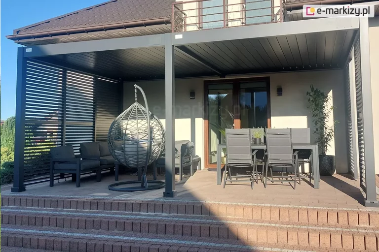 Zdjęcie przedstawia gotową realizację dużej, dwumodułowej pergoli tarasowej zamontowanej przy nowoczesnym domu jednorodzinnym. Konstrukcja wykonana jest z aluminium w kolorze ciemnego antracytu i przylega bezpośrednio do jasnej elewacji budynku, pod balkonem z metalową balustradą. Pergola posiada płaski dach z ruchomych lameli, które są w pozycji zamkniętej. Boczne ściany zabudowy zostały wyposażony w system paneli bocznych z poziomymi, ruchomymi lamelami, które tworzą ażurową ścianę osłaniającą strefę wypoczynkową. Pod zadaszeniem zaaranżowano komfortową przestrzeń relaksu, na którą składają się ciemnoszary zestaw mebli modułowych z grubymi poduszkami ustawiony w narożniku, wiszący fotel typu kokon w kolorze szarym zawieszony na własnym stelażu oraz strefa jadalniana z dużym, ciemnym stołem i czterema dopasowanymi kolorystycznie krzesłami o wysokich oparciach. Całość posadowiona jest na podwyższonym tarasie wyłożonym brązową kostką brukową, do którego prowadzą szerokie schody blokowe. W tle, po prawej stronie przy przeszklonych drzwiach tarasowych, stoi wysoka roślina w ozdobnej, czarnej donicy. Scena jest oświetlona mocnym, naturalnym światłem słonecznym, które rzuca wyraźne cienie na podłoże i elewację budynku.
