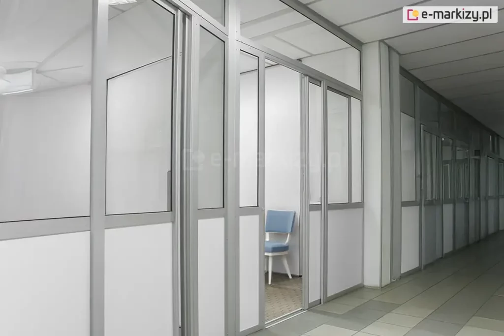 Wewnętrzna zabudowa biurowa wykonana w systemie ścianek działowych Fest, tworząca szereg wydzielonych boksów roboczych wzdłuż korytarza. Konstrukcja opiera się na smukłych profilach aluminiowych w kolorze naturalnego aluminium, które stabilizują wypełnienia szklane oraz nieprzezierne panele. Dolna część modułów oraz wybrane sekcje pionowe zostały wypełnione pełnymi płytami w kolorze białym, natomiast górne partie oraz drzwi przesuwne posiadają wypełnienie z przezroczystego szkła, co zapewnia doświetlenie wydzielonych pomieszczeń. W centralnej części widoczne jest wejście do jednego z boksów z otwartym skrzydłem drzwiowym, ukazujące fragment wnętrza wyposażonego w niebieskie krzesło biurowe na białych nogach oraz szarą wykładzinę dywanową o drobnoziarnistej teksturze. Sufit wewnątrz boksów i na korytarzu jest kasetonowy, w kolorze białym, zintegrowany z systemem oświetlenia technicznego. Posadzka ciągu komunikacyjnego została wyłożona wielkoformatowymi płytami ceramicznymi w dwóch odcieniach szarości, układanymi naprzemiennie w geometryczny wzór. Całość realizacji charakteryzuje się surowym, minimalistycznym designem, typowym dla nowoczesnych przestrzeni biurowych i usługowych