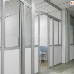 Wewnętrzna zabudowa biurowa wykonana w systemie ścianek działowych Fest, tworząca szereg wydzielonych boksów roboczych wzdłuż korytarza. Konstrukcja opiera się na smukłych profilach aluminiowych w kolorze naturalnego aluminium, które stabilizują wypełnienia szklane oraz nieprzezierne panele. Dolna część modułów oraz wybrane sekcje pionowe zostały wypełnione pełnymi płytami w kolorze białym, natomiast górne partie oraz drzwi przesuwne posiadają wypełnienie z przezroczystego szkła, co zapewnia doświetlenie wydzielonych pomieszczeń. W centralnej części widoczne jest wejście do jednego z boksów z otwartym skrzydłem drzwiowym, ukazujące fragment wnętrza wyposażonego w niebieskie krzesło biurowe na białych nogach oraz szarą wykładzinę dywanową o drobnoziarnistej teksturze. Sufit wewnątrz boksów i na korytarzu jest kasetonowy, w kolorze białym, zintegrowany z systemem oświetlenia technicznego. Posadzka ciągu komunikacyjnego została wyłożona wielkoformatowymi płytami ceramicznymi w dwóch odcieniach szarości, układanymi naprzemiennie w geometryczny wzór. Całość realizacji charakteryzuje się surowym, minimalistycznym designem, typowym dla nowoczesnych przestrzeni biurowych i usługowych