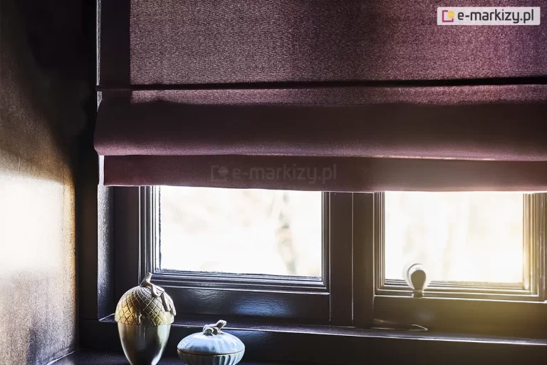 Zdjęcie przedstawia zbliżenie na dolną część rolety rzymskiej w głębokim, śliwkowym lub ciemnofioletowym kolorze, zamontowanej w ciemnej ramie okiennej. Tkanina rolety ma gęsty, satynowy splot z delikatnym połyskiem, który efektownie odbija wpadające światło. Roleta jest częściowo podciągnięta, tworząc charakterystyczne dla tego systemu poziome, miękkie zakładki materiału nachodzące na siebie. Dolna krawędź osłony kończy się w połowie wysokości okna, odsłaniając widok na jasne, rozproszone światło zewnętrzne. Okno posiada ciemne, niemal czarne ramy o klasycznym profilu, z widoczną klamką w dolnej części prawego skrzydła. Na ciemnym parapecie, po lewej stronie, ustawione są dwa przedmioty dekoracyjne: większy pojemnik w kształcie stylizowanego, metalicznego żołędzia o złotawej barwie oraz mniejsza, ceramiczna puzderka w kolorze szarym z ozdobnym detalem na wieczku. Mocne, naturalne światło wpada przez dolną część okna, tworząc wyraźny kontrast między jasnym tłem a nasyconą barwą tkaniny oraz ciemną oprawą stolarki okiennej.