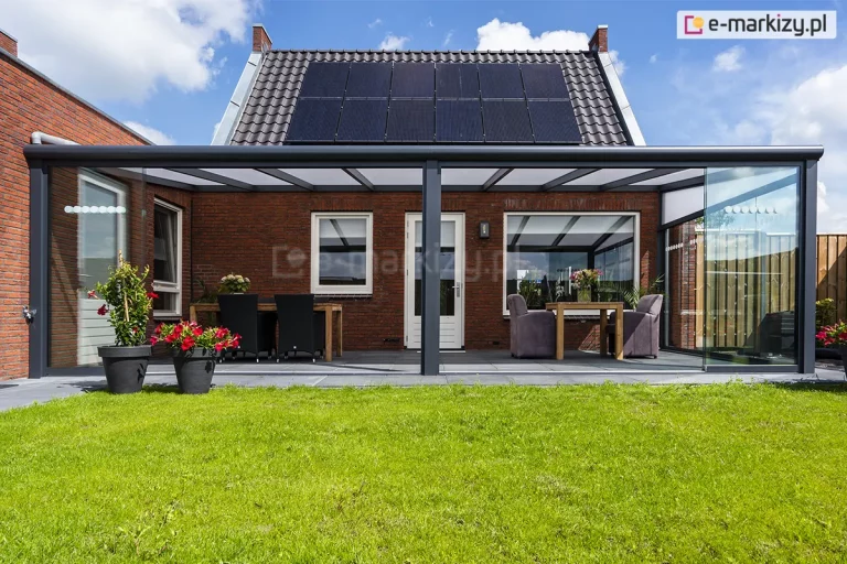 Szerokie, aluminiowe zadaszenie tarasowe w kolorze antracytowym, zamontowane do parterowego budynku wykonanego z ciemnoczerwonej cegły. Konstrukcja zadaszenia posiada dach z płyt poliwęglanowych o mlecznym zabarwieniu, wsparty na systemowych krokwiach i trzech słupach nośnych o kwadratowym przekroju. Boki tarasu zostały zabudowane przesuwnymi panelami szklanymi Auris. Frontowe panele zostały częściowo rozsunięte, otwierając przestrzeń na ogród. Pod zadaszeniem znajdują się dwie oddzielne strefy wypoczynkowe: po lewej stronie ciemny zestaw obiadowy z prostokątnym stołem, a po prawej mniejszy stolik z dwoma tapicerowanymi fotelami w kolorze szarym. Posadzka tarasu wyłożona jest dużymi, ciemnoszarymi płytami, które graniczą z soczyście zielonym trawnikiem. Na pierwszym planie, po lewej stronie, ustawiono dwie czarne donice z kwitnącymi czerwonymi kwiatami. Budynek posiada białą stolarkę okienną i drzwiową, a na dwuspadowym dachu pokrytym ciemną dachówką zainstalowano panele fotowoltaiczne.