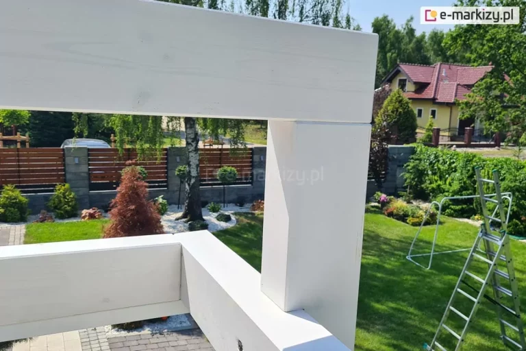 Zbliżenie na miejsce mocowania bramki pergoli drewnianej Moza do jej konstrukcji pomalowanej farbą krylową na biało. W tle widoczny trawnik, na którym stoi drabina i bramka piłkarska oraz ogród z licznymi roślinami, żywopłotem oraz brzozą. W tle widoczny płot z drewnianymi panelami. Po prawej w tle sąsiedni budynek o żółtej fasadzie ze spadzistym dachem pokrytym czerwoną dachówką.
