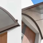 Grafika składa się z dwóch ujęć tego samego daszka naddrzwiowego, prezentujących go z różnych perspektyw na tle jasnej elewacji i błękitnego nieba. Konstrukcja posiada łukowaty kształt i opiera się na ciemnobrązowych profilach aluminiowych. Wypełnienie zadaszenia wykonane jest z poliwęglanu komorowego o dymionym, brązowym zabarwieniu, przez który prześwituje charakterystyczna struktura pionowych kanalików. Lewe zdjęcie ukazuje daszek z perspektywy bocznej, patrząc od dołu. Widoczna jest precyzyjnie wykończona krawędź boczna oraz przedni profil rynnowy, który domyka taflę poliwęglanu. Na górnej powierzchni profilu, przy krawędzi przyściennej, zamontowany jest niewielki, ciemny uchwyt montażowy. Pod zadaszeniem widoczny jest fragment brązowych drzwi. Prawe zdjęcie przedstawia daszek z perspektywy dolnej, co pozwala zaobserwować spód konstrukcji. Przez dymiony poliwęglan przebiega poprzeczna, cienka listwa wzmacniająca, która stabilizuje łukowate wygięcie płyty. Widoczny jest sposób osadzenia wypełnienia w bocznych prowadnicach ramy. W dolnej części kadru widać górną krawędź wnęki drzwiowej oraz fragment skrzydła drzwiowego w kolorze naturalnego drewna. Całość kompozycji na prawym zdjęciu dopełnia jasne, czyste niebo widoczne w tle za krawędzią daszka.