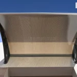 Zdjęcie prezentuje widok od dołu na zamontowany daszek drzwiowy o nowoczesnej, wygiętej konstrukcji, zainstalowany nad wnęką wejściową na tle jasnej elewacji i błękitnego nieba. Wypełnienie zadaszenia stanowi płyta poliwęglanowa w kolorze dymnego brązu o wyraźnej, pionowej strukturze komorowej, która jest ujęta w srebrne profile wykończeniowe na krawędzi przyściennej oraz czołowej. Całość konstrukcji opiera się na dwóch solidnych, czarnych wspornikach bocznych o ażurowym wzornictwie z geometrycznymi wzmocnieniami, które są przytwierdzone bezpośrednio do górnej krawędzi muru. Perspektywa zdjęcia podkreśla szerokość zadaszenia oraz sposób, w jaki łukowato wygięta połaci osłania przestrzeń przed wejściem, zapewniając ochronę przed opadami atmosferycznymi przy zachowaniu wysokiej estetyki wykończenia detali montażowych.