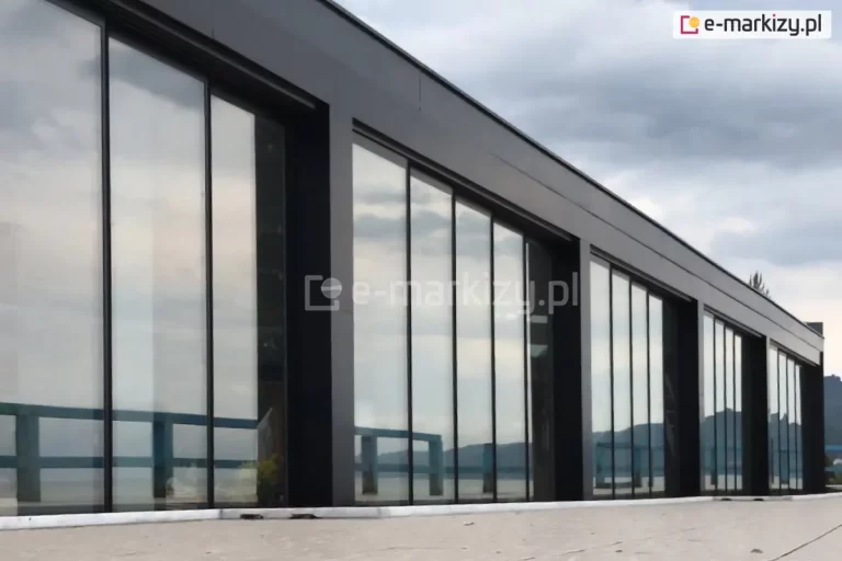 Nowoczesna elewacja budynku z zabudową wykonaną z systemu bezramowych szklanych ścian przesuwnych Miros, osadzonych w aluminiowej konstrukcji w kolorze czarnym. Przeszklone panele o dużej wysokości tworzą jednolitą taflę, odbijającą niebo, linię horyzontu i otoczenie. Konstrukcja oparta jest na rytmicznie rozmieszczonych, pionowych profilach aluminiowych, które dzielą powierzchnię fasady na symetryczne segmenty. Na dole widoczna niska listwa prowadząca oraz fragment posadzki wyłożonej jasnym materiałem. W tle za przeszkleniem widać niebieską balustradę, jezioro lub zbiornik wodny oraz delikatnie zarysowane górskie wzniesienia. Nad całością niebo z zachmurzeniem i przebijającym się światłem.