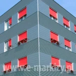 Nowoczesna bryła budynku biurowego o charakterystycznej elewacji wykonanej z szarej, ryflowanej blachy trapezowej, która tworzy regularny, poziomy wzór tekstury. Architektura obiektu została wzbogacona o system zewnętrznych osłon przeciwsłonecznych markizoleta z tkaniną typu screen w intensywnym, czerwonym kolorze, które zainstalowano w otworach okiennych na wszystkich widocznych kondygnacjach. Każda z rolet posiada mechanizm wysięgnikowy, pozwalający na uchylenie dolnej części tkaniny w celu regulacji stopnia zaciemnienia pomieszczeń przy jednoczesnym zachowaniu dostępu do naturalnego światła i wentylacji. Widok pod kątem ukazuje narożnik budynku, na którym symetrycznie rozmieszczono rzędy okien z opuszczonymi przesłonami, tworzącymi wyraźny akcent kolorystyczny na tle chłodnej, industrialnej elewacji. Konstrukcja osłon jest zintegrowana z ramami okiennymi, a precyzyjne ustawienie wysięgników nadaje fasadzie dynamicznego i uporządkowanego wyglądu, typowego dla nowoczesnych projektów budownictwa komercyjnego. Bezchmurne, jasnoniebieskie niebo w tle potęguje kontrast między ciepłą barwą tkanin a surową kolorystyką metalowych płyt ściennych.