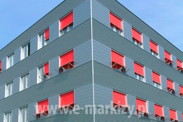 Nowoczesna bryła budynku biurowego o charakterystycznej elewacji wykonanej z szarej, ryflowanej blachy trapezowej, która tworzy regularny, poziomy wzór tekstury. Architektura obiektu została wzbogacona o system zewnętrznych osłon przeciwsłonecznych markizoleta z tkaniną typu screen w intensywnym, czerwonym kolorze, które zainstalowano w otworach okiennych na wszystkich widocznych kondygnacjach. Każda z rolet posiada mechanizm wysięgnikowy, pozwalający na uchylenie dolnej części tkaniny w celu regulacji stopnia zaciemnienia pomieszczeń przy jednoczesnym zachowaniu dostępu do naturalnego światła i wentylacji. Widok pod kątem ukazuje narożnik budynku, na którym symetrycznie rozmieszczono rzędy okien z opuszczonymi przesłonami, tworzącymi wyraźny akcent kolorystyczny na tle chłodnej, industrialnej elewacji. Konstrukcja osłon jest zintegrowana z ramami okiennymi, a precyzyjne ustawienie wysięgników nadaje fasadzie dynamicznego i uporządkowanego wyglądu, typowego dla nowoczesnych projektów budownictwa komercyjnego. Bezchmurne, jasnoniebieskie niebo w tle potęguje kontrast między ciepłą barwą tkanin a surową kolorystyką metalowych płyt ściennych.